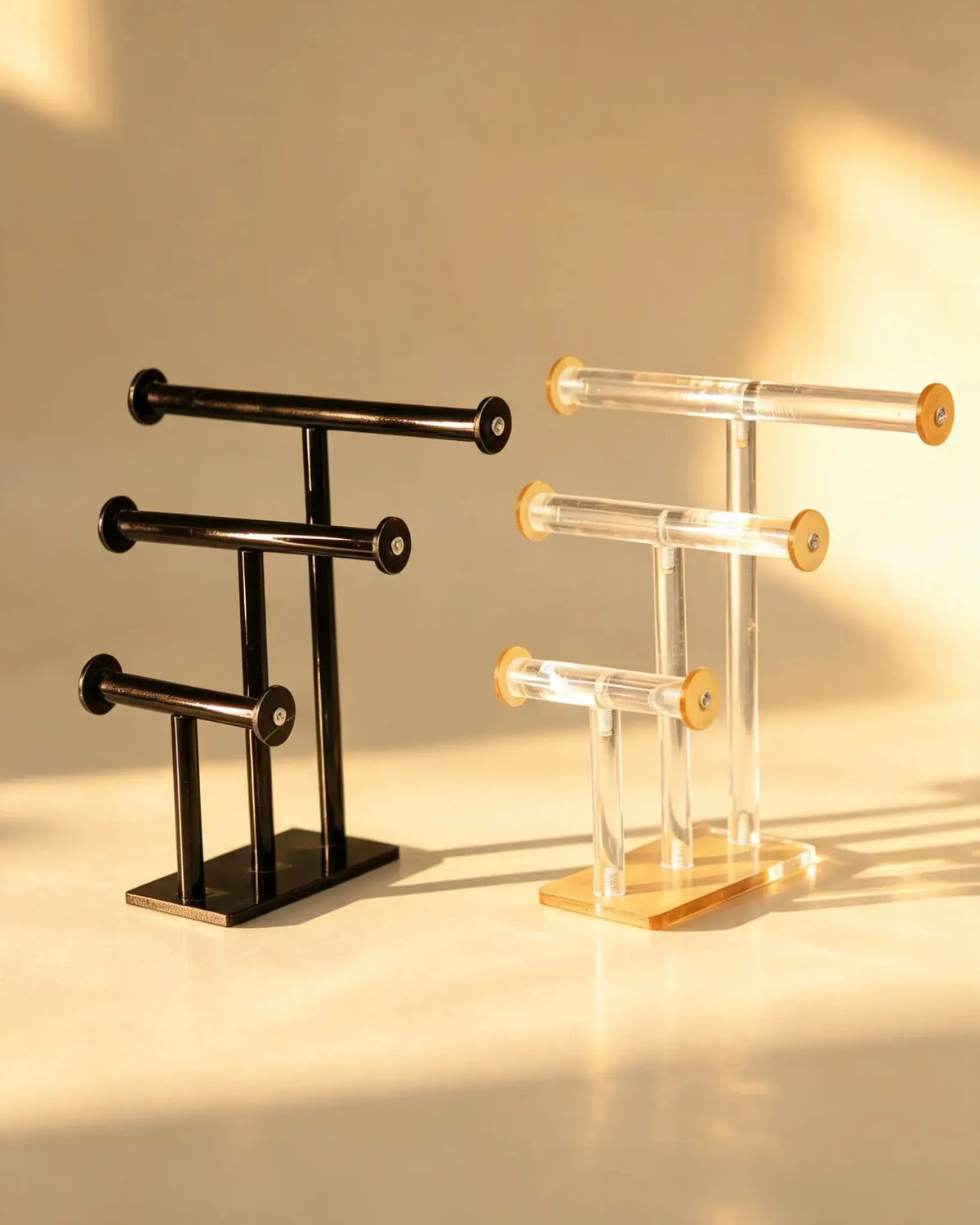 Two three-tier T-bar bangle stands side by side, one black metal and one clear acrylic with gold-tone caps