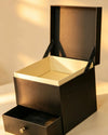 Black gift box with hinged lid open, showing cream-lined interior and a pull-out drawer with a knob