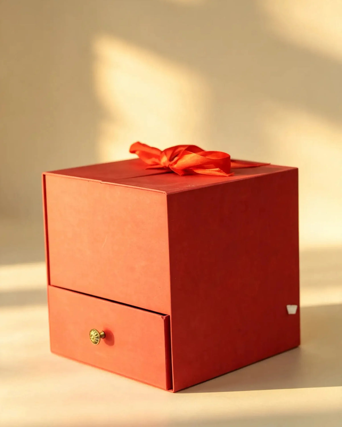 Terracotta red cube gift box with a pull-out drawer, brass knob, and ribbon bow on the lid