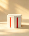 White hexagon gift box closed, with a red ribbon stripe down the front, on a warm beige backdrop