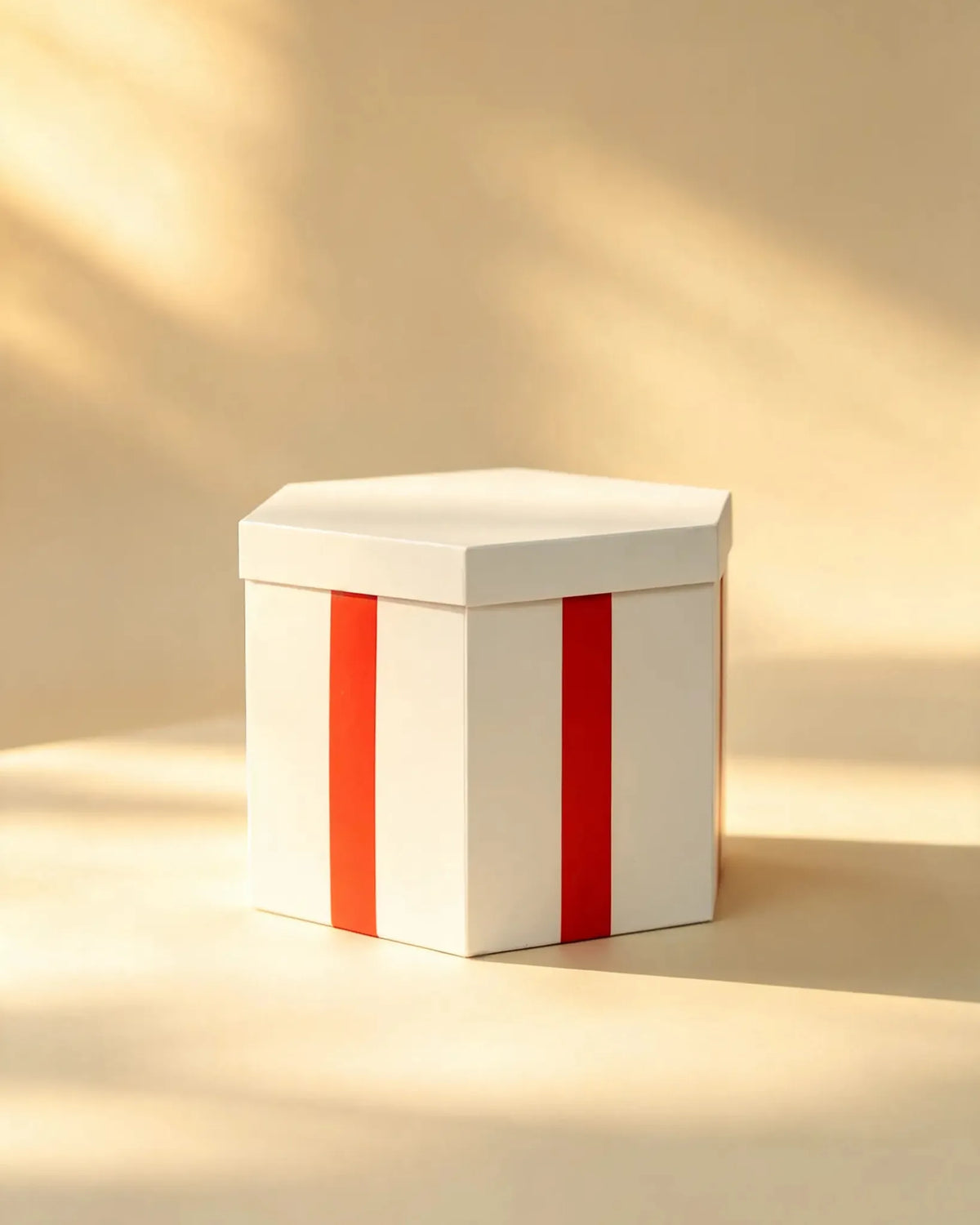 White hexagon gift box closed, with a red ribbon stripe down the front, on a warm beige backdrop