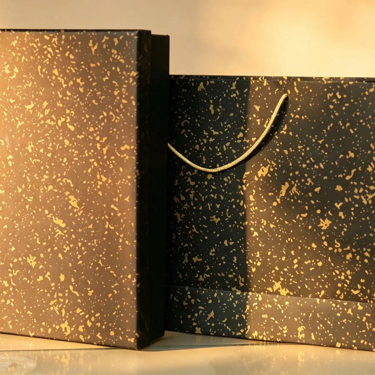 Black gift box with gold speckle print beside a matching carry bag with gold cord handle
