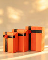 Three nesting orange gift boxes with lids in graduating sizes, each tied with a black ribbon and red flower