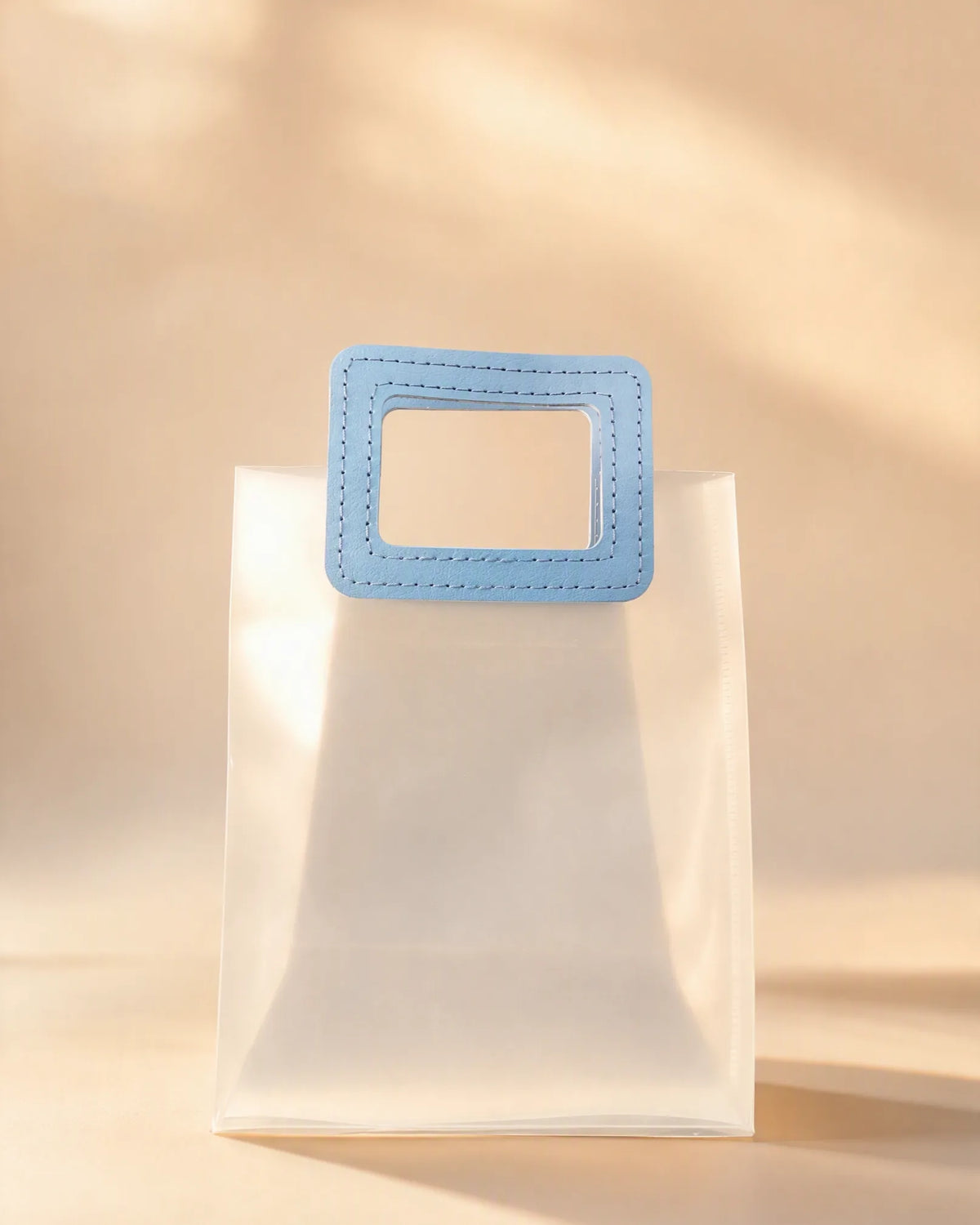 Clear PVC gift bag with blue square handle against a warm beige backdrop