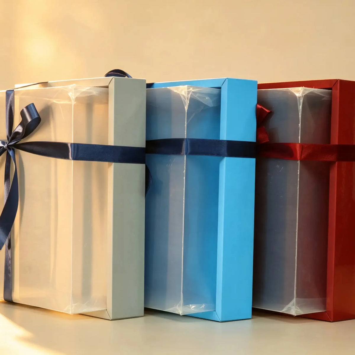 Clear-window PVC gift boxes in cream, sky blue and deep red with satin ribbon bows, shown together