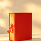 Red rigid gift box with gold-tone interior and orange ribbon bow, closed