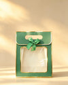 Green paper gift bag with gold foil trim, clear front window, and a green ribbon bow on the handle