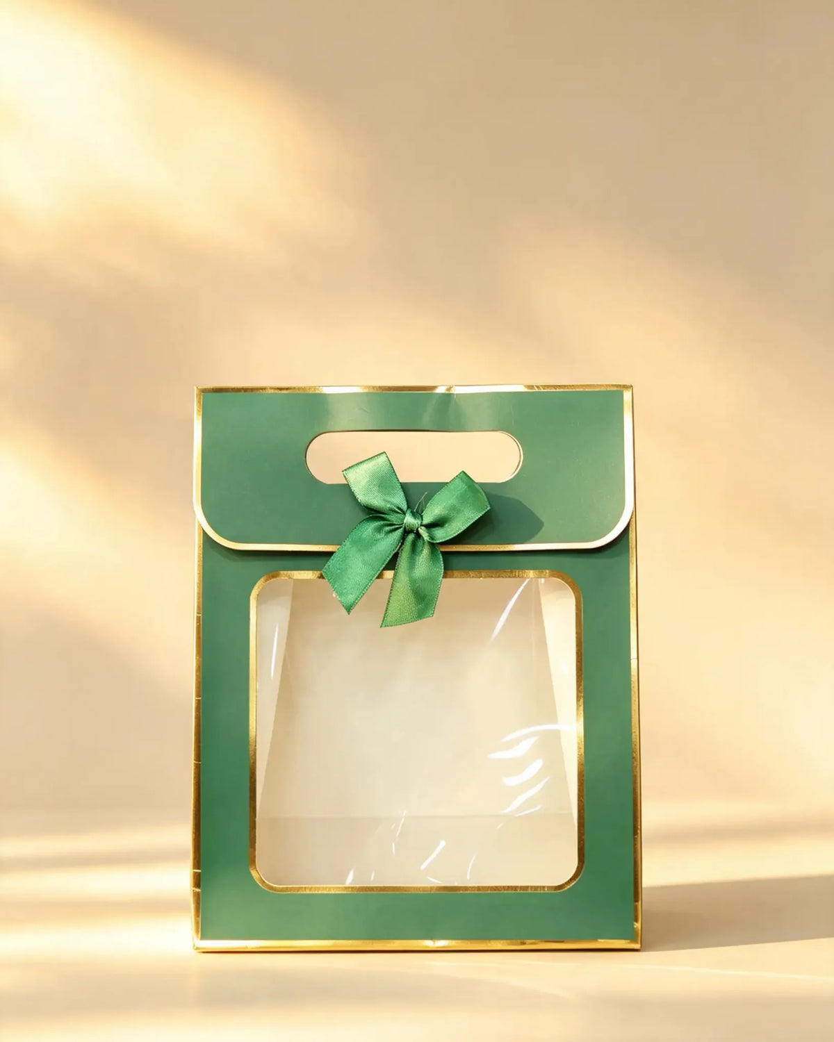Green paper gift bag with gold foil trim, clear front window, and a green ribbon bow on the handle