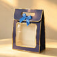 Navy blue gift bag with gold foil trim, clear front window, and a blue ribbon bow, angled view